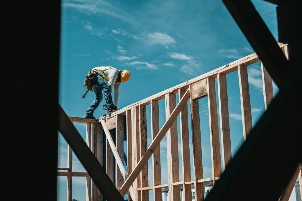 Ein Mann in Arbeitskleidung mit Schutzhelm und Werkzeug arbeitet auf einem Holzgerüst vor blauem Himmel